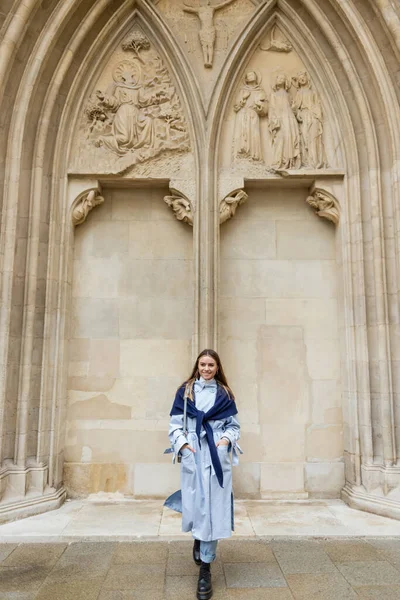 Happy woman with scarf on top of blue trench coat standing with hands in pockets near historical building in Vienna — Stock Photo