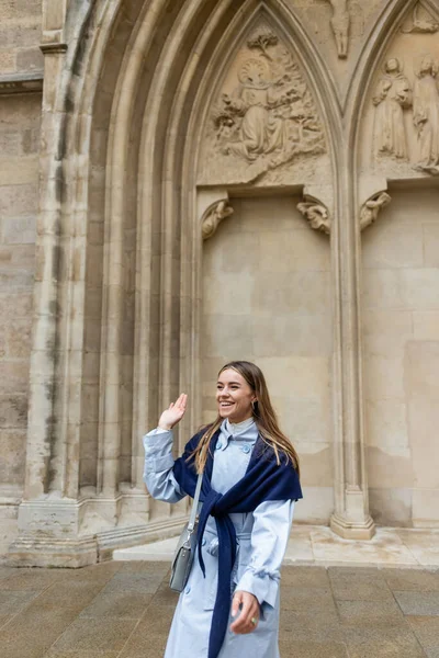Joyeuse jeune femme avec écharpe sur le dessus du trench coat bleu agitant la main près du bâtiment historique à Vienne — Photo de stock