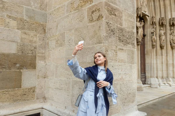 Junge Frau mit Schal über blauem Trenchcoat macht Selfie in der Nähe historischer Gebäude in Wien — Stockfoto