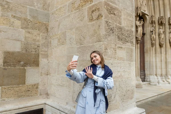 Щаслива жінка з шарфом на синьому плащі має відеодзвінок біля історичного будинку у Відні. — стокове фото