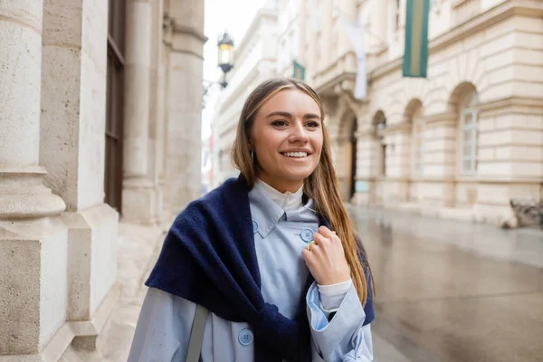 Mujer feliz y elegante con bufanda en la parte superior de la gabardina azul sonriendo mientras mira hacia otro lado en la calle en Viena - foto de stock