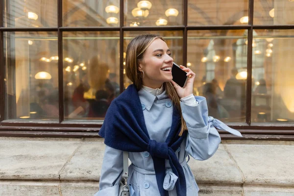 Веселая женщина с шарфом на голубом пальто разговаривает по смартфону на улице в Вене — стоковое фото