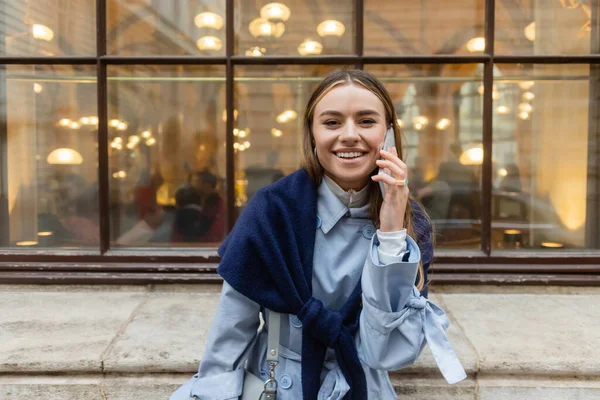 Mujer sonriente con bufanda en la parte superior de la gabardina azul hablando en el teléfono inteligente en la calle en Viena - foto de stock