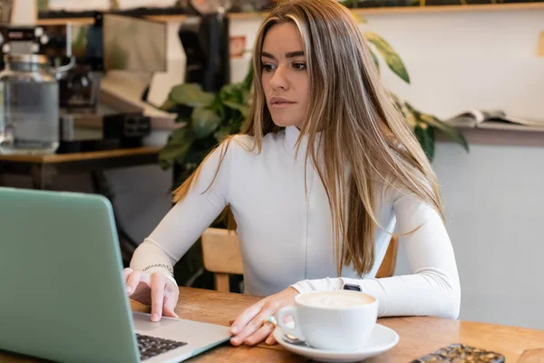 Mulher encantadora em gola alta usando laptop perto de xícara de cappuccino no café — Fotografia de Stock