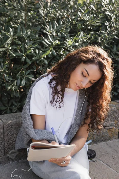 Young and curly woman in casual clothes listening music in wired earphones and writing on notebook while relaxing on stone bench in park in Barcelona, Spain — Stock Photo