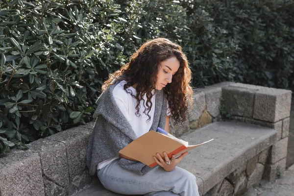 Jeune femme brune bouclée en t-shirt et pull écrivant sur un carnet tout en passant du temps sur un banc de pierre près de buissons verts dans un parc pendant la journée à Barcelone, Espagne — Photo de stock