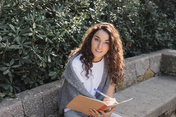 Carefree young and curly woman in sweater and t-shirt looking at camera while writing on notebook and spending time on bench near green bushes in park in Barcelona, Spain — Stock Photo