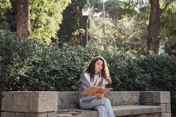 Mulher jovem e encaracolado Pensivo em roupas casuais segurando notebook e marcador perto de lábios enquanto sentado perto de dispositivos no banco de pedra e plantas verdes no parque durante o dia em Barcelona, Espanha — Fotografia de Stock