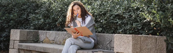 Сфокусована молода кучерява жінка дивиться на камеру, тримаючи маркер і зошит біля смартфона і ноутбука на кам'яній лавці біля кущів парку в Барселоні, Іспанія, банер, робота з будь-якого місця — стокове фото