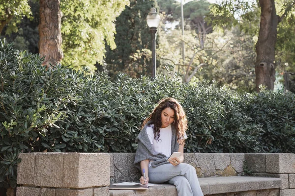 Joven y rizado freelancer en ropa casual usando un smartphone y escribiendo en un cuaderno mientras está sentado cerca de un portátil en un banco de piedra y arbustos verdes en el parque en Barcelona, España - foto de stock