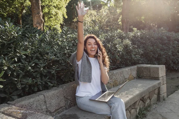 Mujer alegre y rizada saludando con la mano y hablando en el teléfono inteligente, mirando hacia otro lado y sosteniendo el ordenador portátil mientras está sentado en el banco de piedra en el parque verde durante el día en Barcelona, España - foto de stock