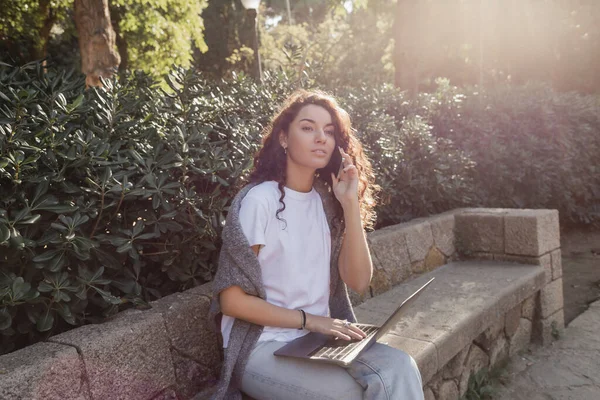 Young and curly freelancer in casual clothes talking on smartphone and looking away while using laptop and sitting on stone bench in green park in Barcelona, Spain — Stock Photo