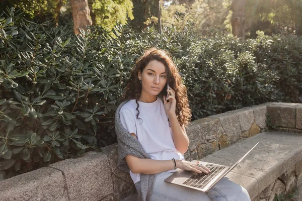 Young and curly freelancer in casual clothes talking on smartphone and using laptop while looking at camera on stone bench near green bushes in park in Barcelona, Spain — Stock Photo
