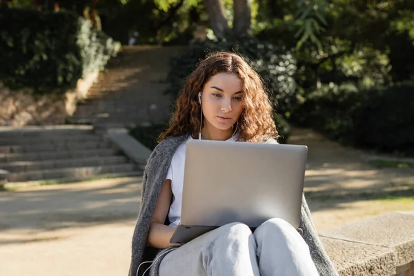 Young and curly freelancer in wired earphones using laptop while sitting on parapet and spending time in blurred green park at daytime in Barcelona, Spain — Stock Photo