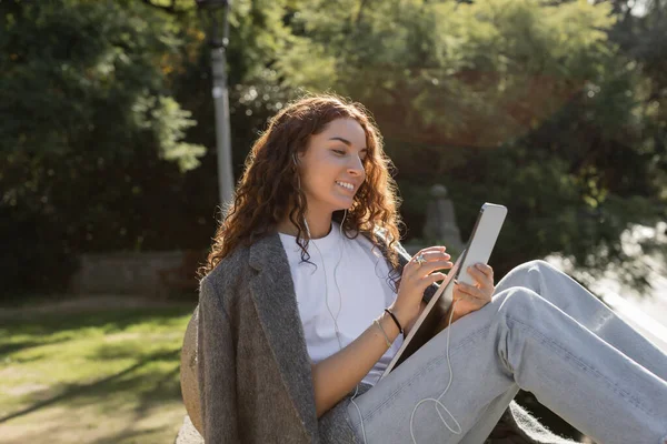 Carefree young and curly freelancer in wired earphones using smartphone and holding laptop while spending time in blurred green park at daytime in Barcelona, Spain — Stock Photo