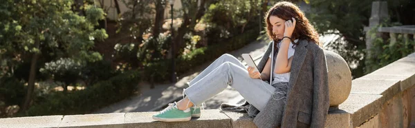 Дивний молодий фрілансер у теплому піджаку з використанням смартфона з навушниками і ноутбука, сидячи на кам'яній лавці в розмитому парку в Барселоні (Іспанія). — стокове фото