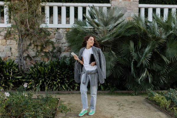 Mujer joven y rizada positiva en auriculares y chaqueta cálida sosteniendo gadgets y mirando hacia otro lado mientras camina en el parque cerca de palmeras durante el día en Barcelona, España - foto de stock