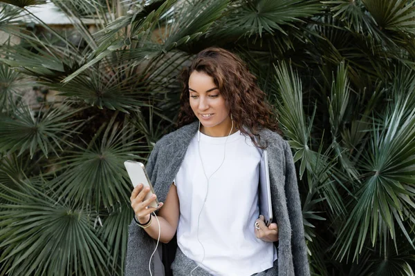 Cheerful young and curly freelancer in warm jacket using earphones and smartphone while holding laptop and standing near green palm trees in park in Barcelona, Spain — Stock Photo