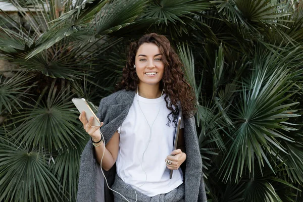 Young and curly freelancer wearing warm jacket and using earphones while holding smartphone and notebook, smiling at camera and standing near palm trees in park in Barcelona, Spain — Stock Photo