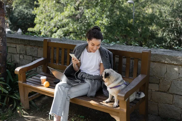 Smiling freelancer in earphones holding smartphone and looking at pug dog while sitting near laptop, coffee to go and orange on wooden bench and spending time in park in Barcelona, Spain — Stock Photo