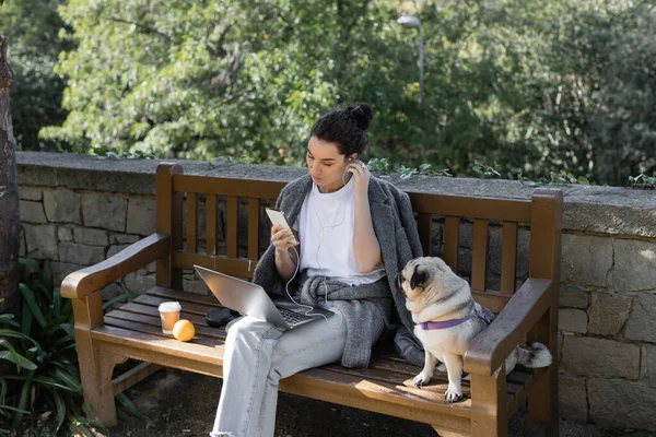 Junge und lockige Freiberuflerin hört Musik mit Kopfhörern, nutzt Smartphone in der Nähe von Laptop und sitzt neben Mops Hund, Orange und Kaffee auf Holzbank im Park in Barcelona, Spanien — Stockfoto