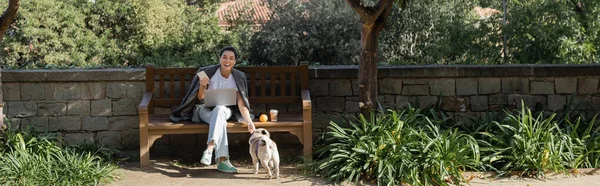 Cheerful young freelancer in earphones holding smartphone and petting pug dog while sitting near laptop, coffee to go and orange on wooden bench in park in Barcelona, Spain, banner, orange tree — Stock Photo