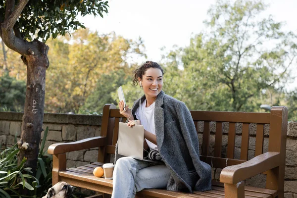 Smiling brunette freelancer in warm jacket using earphones and devices while spending time near pug dog, coffee to go and orange on wooden bench in park in Barcelona, Spain — Stock Photo