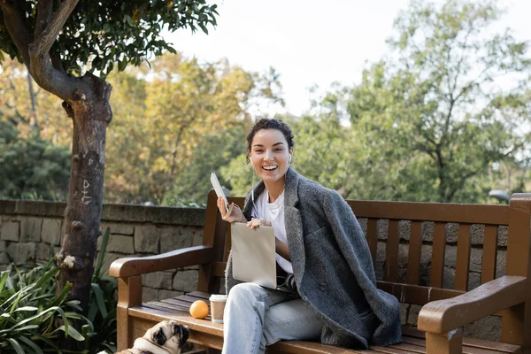 Cheerful young freelancer in warm jacket listening music in earphones, looking at camera and using devices near pug dog and takeaway coffee on wooden bench in park in Barcelona, Spain — Stock Photo
