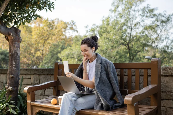 Smiling young freelancer in warm jacket and earphones smiling, covering mouth, having video call on smartphone near laptop and coffee to go on wooden bench in park in Barcelona, Spain — Stock Photo