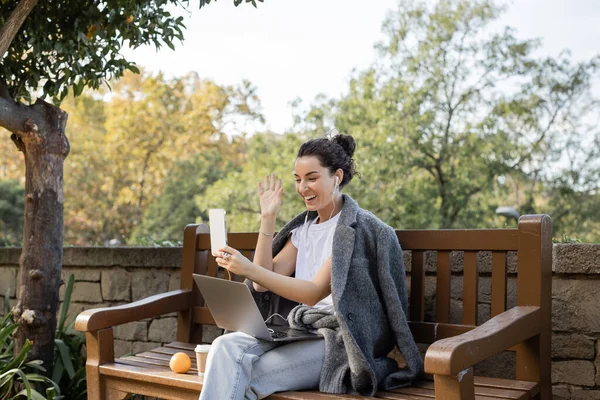 Cheerful young freelancer in warm jacket and earphones waving hand at smartphone while having video call on smartphone near laptop, coffee to go and orange on wooden bench in park in Barcelona, Spain — Stock Photo