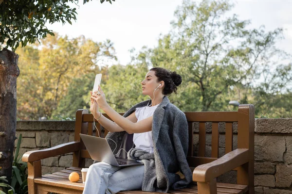 Side view of young freelancer in warm jacket using earphones while having video chat on smartphone near laptop, coffee to go and orange on wooden bench in park in Barcelona, Spain — Stock Photo