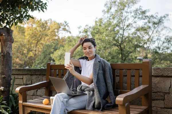 Smiling and young freelancer in warm jacket using earphones and smartphone while sitting near laptop, takeaway drink and fresh orange on wooden bench in park in Barcelona, Spain — Stock Photo