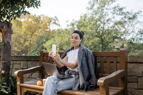 Joven freelancer despreocupado en auriculares usando teléfono celular y sosteniendo el portátil mientras está sentado cerca de naranja y café para ir a un banco de madera en el parque en Barcelona, España - foto de stock
