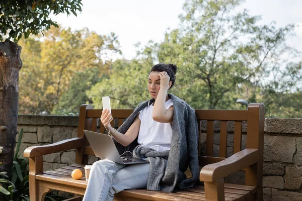 Riccio e grazioso freelance in giacca calda con videochiamata su smartphone vicino a laptop, caffè per andare e arancione su una panchina di legno nel parco di giorno a Barcellona, Spagna — Foto stock