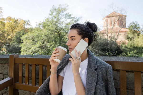 Mujer bonita y rizada en camiseta y chaqueta cálida hablando en smartphone y sosteniendo café para llevar sentado en un banco de madera en el parque durante el día en Barcelona, España, taza de papel con bebida caliente - foto de stock