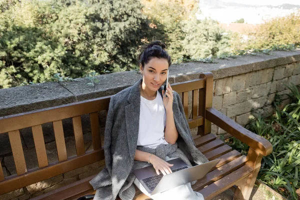 Lächelnde junge Frau in warmer Jacke, die mit dem Smartphone spricht und in die Kamera schaut, während sie am Laptop arbeitet und auf einer Holzbank im Park in Barcelona sitzt, Spanien, Arbeit von überall — Stockfoto