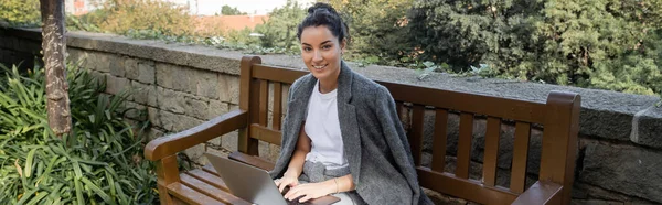 Cheerful young brunette freelancer in warm jacket looking at camera while working on laptop and sitting on wooden bench in park at daytime in Barcelona, Spain, banner — Stock Photo