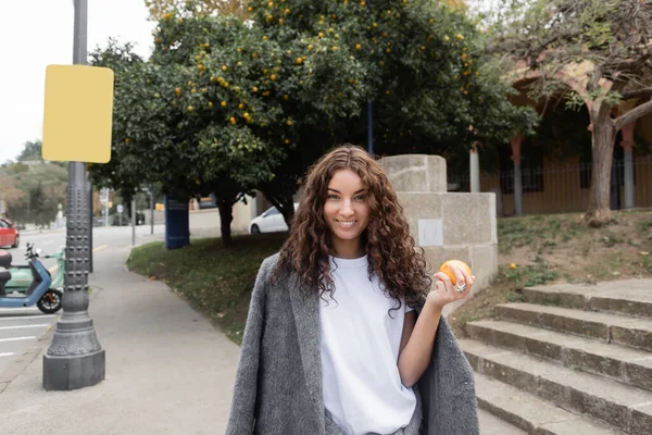 Femme souriante jeune et bouclée en veste chaude tenant orange frais et regardant la caméra tout en se tenant debout sur la rue urbaine floue à l'arrière-plan à Barcelone, Espagne, lampadaire, scooter moteur — Photo de stock