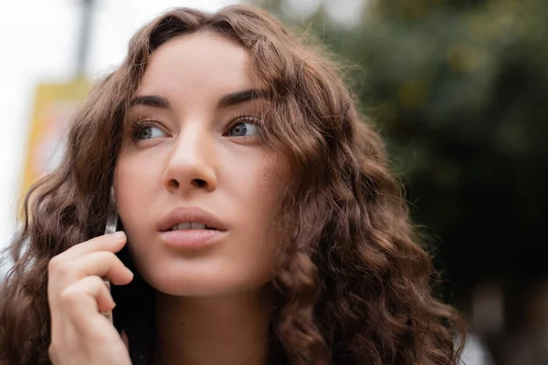 Portrait of young brunette and curly woman looking away while having conversation on smartphone and standing on blurred urban street in Barcelona, Spain — Stock Photo