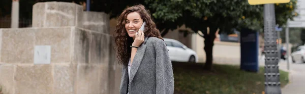 Positive young and curly brunette woman in casual jacket looking at camera and talking on cellphone while standing on blurred urban street in Barcelona, Spain, banner — Stock Photo