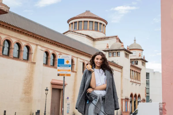 Smiling and brunette woman in casual jacket holding fresh orange and leash while standing near blurred historic landmark on urban street in Barcelona, Spain, ancient building — Stock Photo