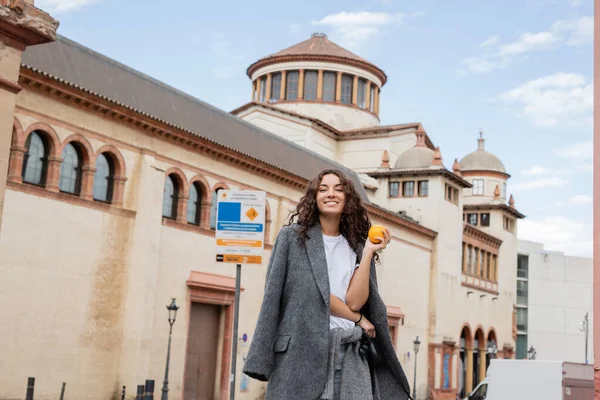 Cheerful young and curly woman in casual warm jacket holding ripe orange and leash while looking at camera near blurred historical landmark on urban street in Barcelona, Spain, ancient building — Stock Photo