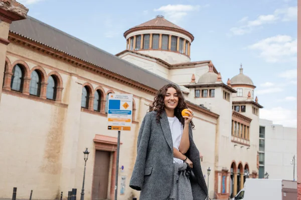 Positive junge Frau in lässiger grauer Jacke mit frischem Orange und Leine, während sie in die Kamera mit historischem Wahrzeichen auf der Stadtstraße in Barcelona blickt, Spanien, antikes Gebäude — Stockfoto