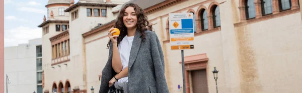 Mujer rizada joven en chaqueta gris casual sosteniendo naranja madura y fresca y sonriendo a la cámara con hito histórico en el fondo al aire libre en Barcelona, España, pancarta, edificio antiguo - foto de stock
