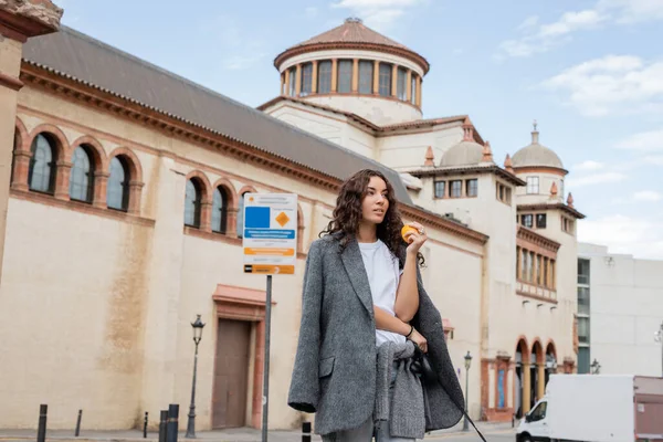 Young curly woman in grey casual jacket holding fresh orange and leash while looking away with blurred historical landmark at background in Barcelona, Spain, ancient building — Stock Photo