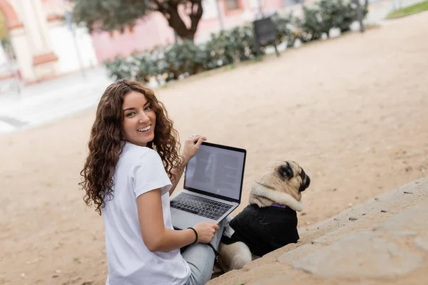 Веселая молодая кудрявая женщина в повседневной одежде смотрит в камеру, держа ноутбук рядом с собакой-мопсом на лестнице в размытом парке Барселоны, Испания, белая футболка — стоковое фото