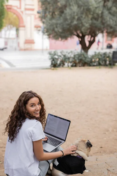 Unbekümmerte junge und lockige Freiberuflerin in lässiger Kleidung, die in die Kamera schaut, während sie Laptop und Streichelmops auf der Treppe im verschwommenen Park in Barcelona benutzt, weißes T-Shirt — Stockfoto