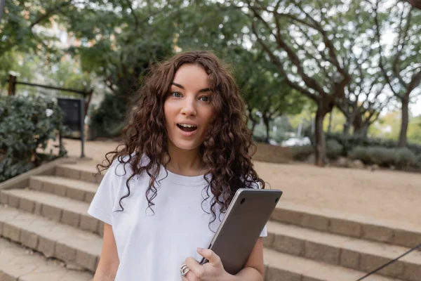 Shocked young and curly freelancer in white t-shirt looking at camera with open mouth while holding laptop and spending time in blurred park at daytime in Barcelona, Spain, — Stock Photo