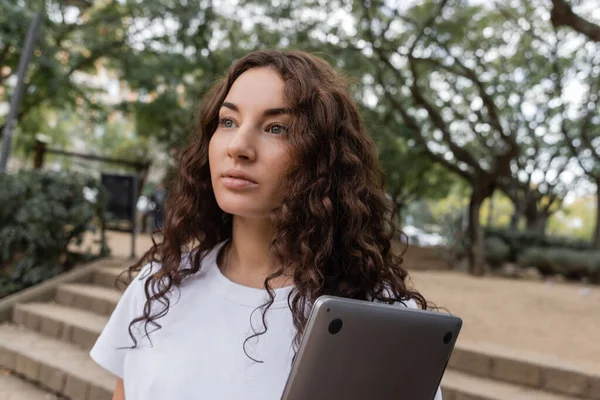 Ritratto di giovane donna bruna in t-shirt bianca che tiene il computer portatile e distoglie lo sguardo mentre trascorre del tempo nel parco sfocato di Barcellona, Spagna — Foto stock