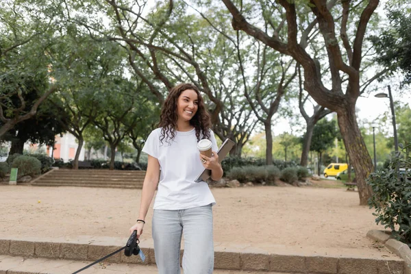 Freelancer jovem e encaracolado sorridente em roupas casuais segurando café para ir, laptop e trela enquanto caminha no parque turvo durante o dia em Barcelona, Espanha — Fotografia de Stock